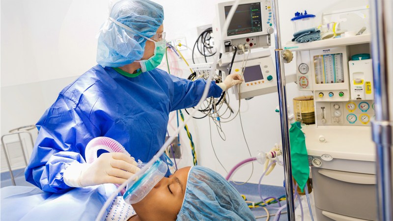 An anesthesiologist in blue scrubs and face mask applies anesthesia to a patient while looking at her monitors.