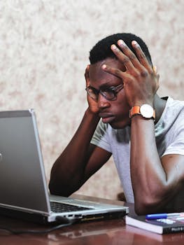 A man appears frustrated while working on a laptop in an office setting, showcasing remote work challenges.