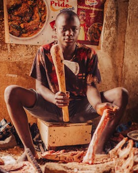 A young butcher sitting indoors, preparing meat with a cleaver, showing traditional techniques.